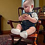 baby, child, denim_overalls, wooden_chair, rocking_chair, toy, indoor, carpet, books, furniture, soft_light, curtains, floor, person, cute, socks, portrait, seated, room, thinking