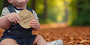Liam is registered to the contest to win money with this photo: baby, child, seven_months, autumn, leaves, forest, denim_overalls, white_shoes, smiling, sitting, nature, outdoor, happy, portrait, cute, young_child, holding_sign, seasonal, fall, baby_clothing