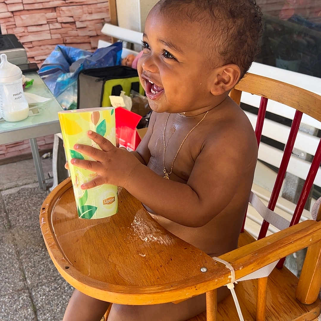 Raphaël a rejoint le concours — aidez-le/la à gagner de superbes lots ! baby, background_objects, bare_chested, child, cup, curly_hair, daylight, drink, footwear_absent, happy, high_chair, necklace, outdoor, person, plastic_bottle, playful, smiling, table, toddler, wooden_furniture