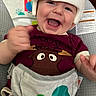 baby, helmet, smiling, laughing, infant, baby_seat, cushion, maroon_shirt, cartoon_moose, colorful, rainbow, sun_illustration, cute, happy, child, playful, indoor, closeup, clothing, protection