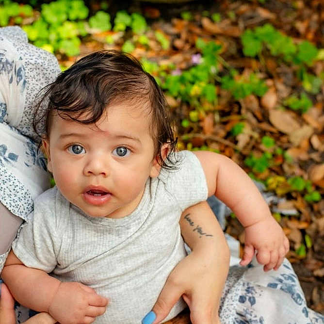 Liam joined the competition — help win amazing prizes! adult, arm, baby, blue_nail_polish, child, curly_hair, expression, gray_shirt, greenery, hand, holding, infant, leaves, looking_up, nails, nature, outdoor, person, skin, tattoo