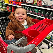 Kai is registered to the contest to win money with this photo: baby, shopping_cart, grocery_store, coffee, smiling, child, clothing, jacket, red_cart, aisle, socks, pacifier, metal_cart, person, retail, shopping, indoor, happy, infant, seat