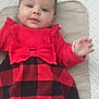 baby, child, infant, dress, red_clothing, black_clothing, plaid, bow, cushion, textured_background, hand, face, eyes, lying_down, cute, indoor, person, smiling, soft_lighting, young_child