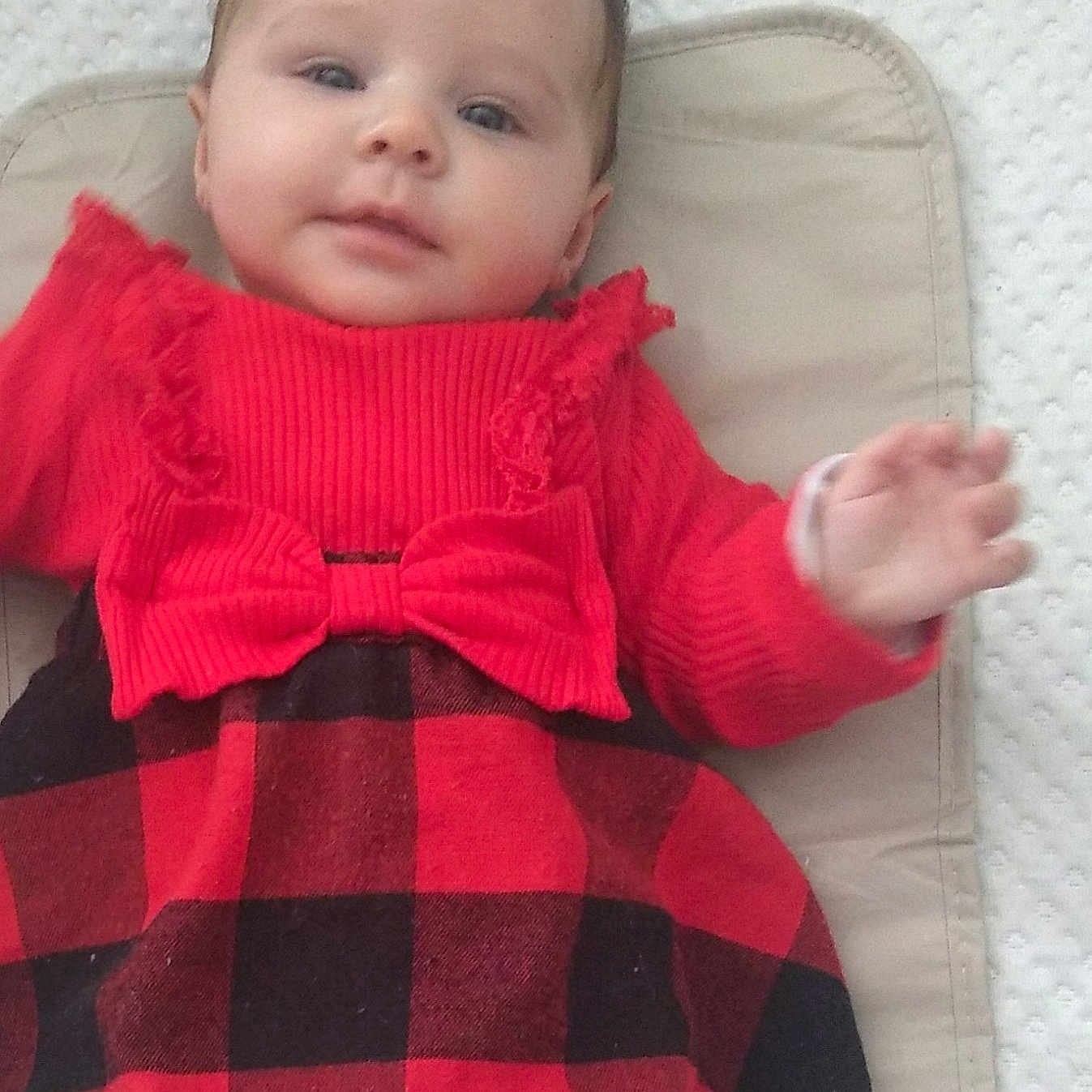 Lynné is registered to the contest to win money with this photo: baby, black_clothing, bow, child, cushion, cute, dress, eyes, face, hand, indoor, infant, lying_down, person, plaid, red_clothing, smiling, soft_lighting, textured_background, young_child