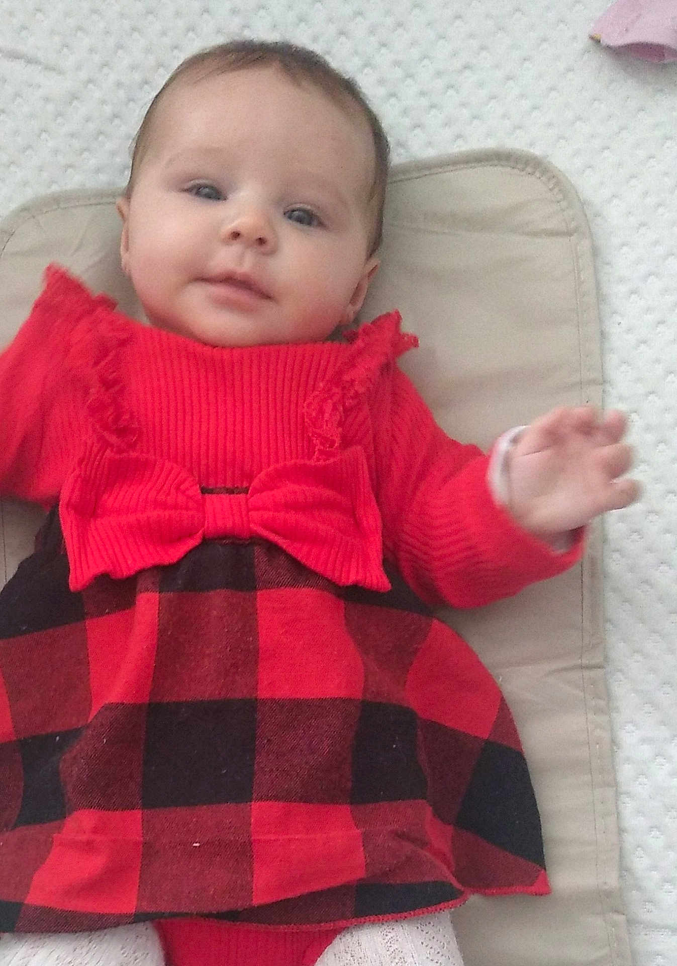 Lynné is registered to the contest to win money with this photo: baby, child, infant, dress, red_clothing, black_clothing, plaid, bow, cushion, textured_background, hand, face, eyes, lying_down, cute, indoor, person, smiling, soft_lighting, young_child