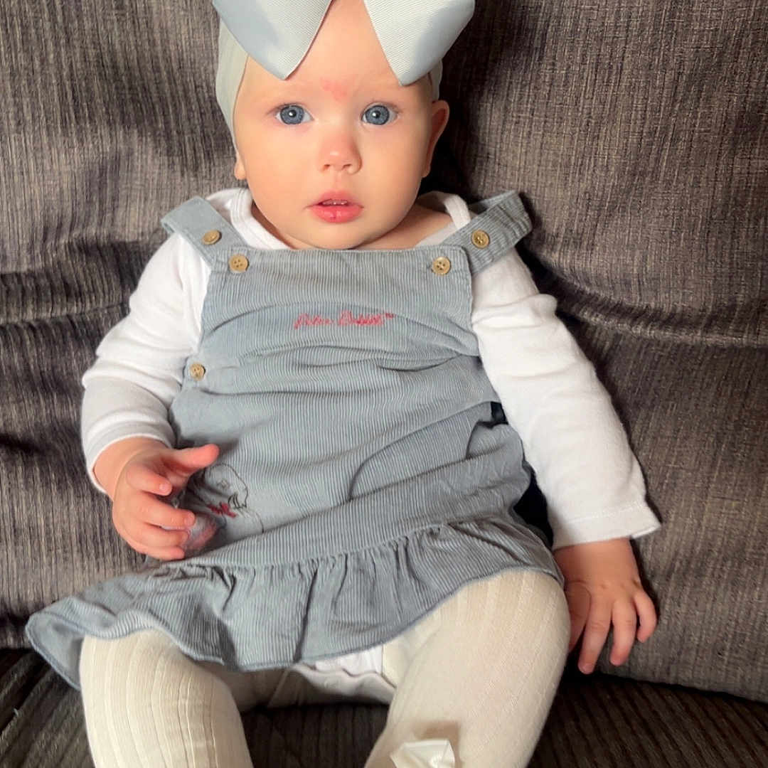 Orla is registered to the contest to win money with this photo: baby, blue_eyes, child, clothing, couch, cute, dress, fashion, gray_couch, headwear, indoor, infant, long_sleeve_shirt, person, plush, portrait, sitting, tights, white_bow, young_child