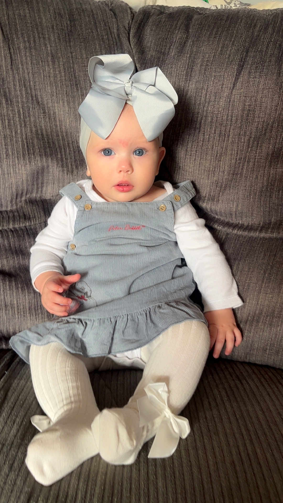 Orla is registered to the contest to win money with this photo: baby, child, blue_eyes, white_bow, dress, tights, sitting, couch, gray_couch, plush, long_sleeve_shirt, indoor, cute, portrait, infant, fashion, clothing, person, young_child, headwear