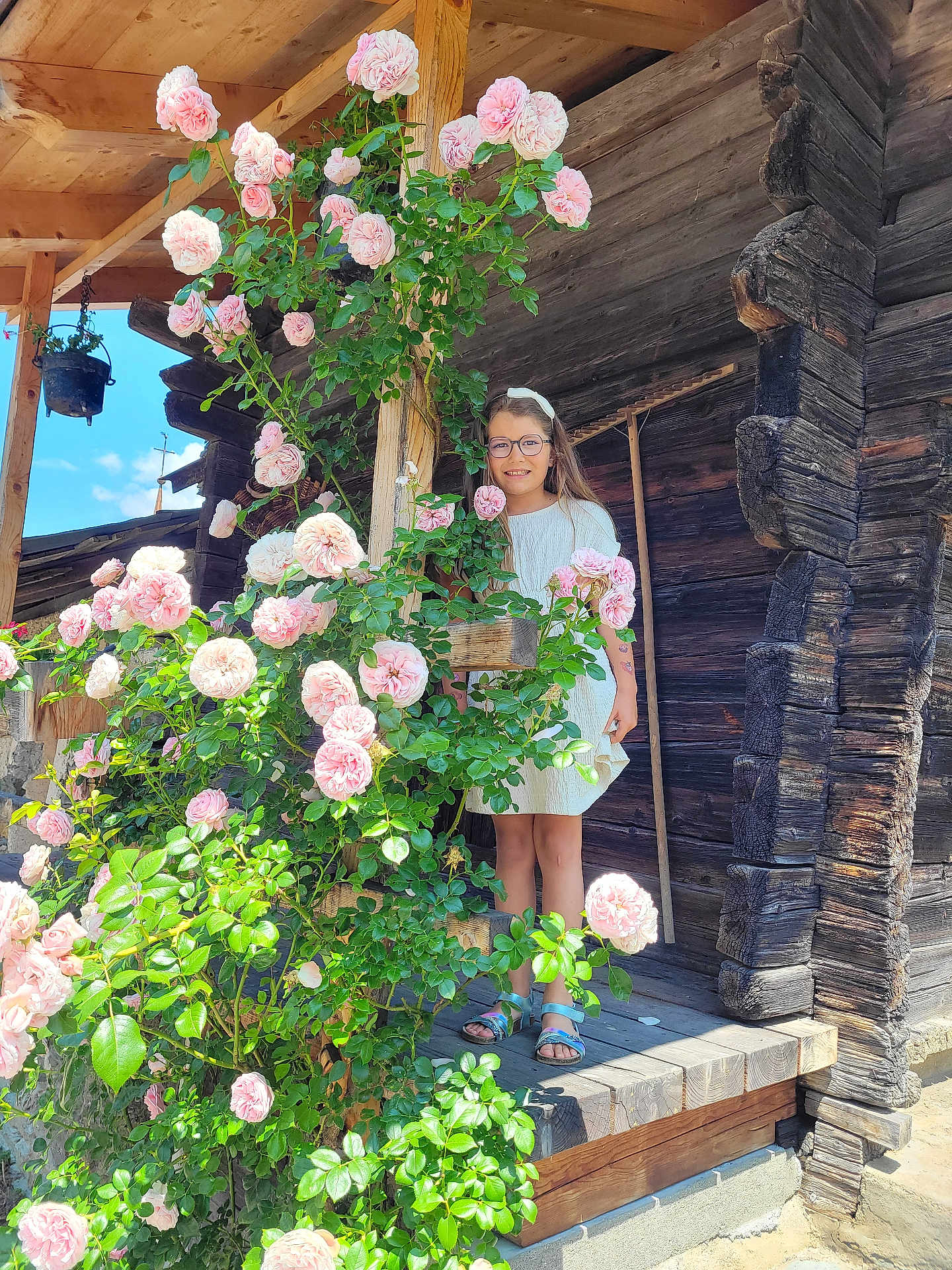 Agathe participe au concours pour gagner de l'argent avec cette photo : child, girl, flowers, roses, glasses, white_dress, wooden_porch, log_cabin, greenery, plants, outdoor, sunlight, nature, smiling, summer, leaves, wood, standing, happy, portrait