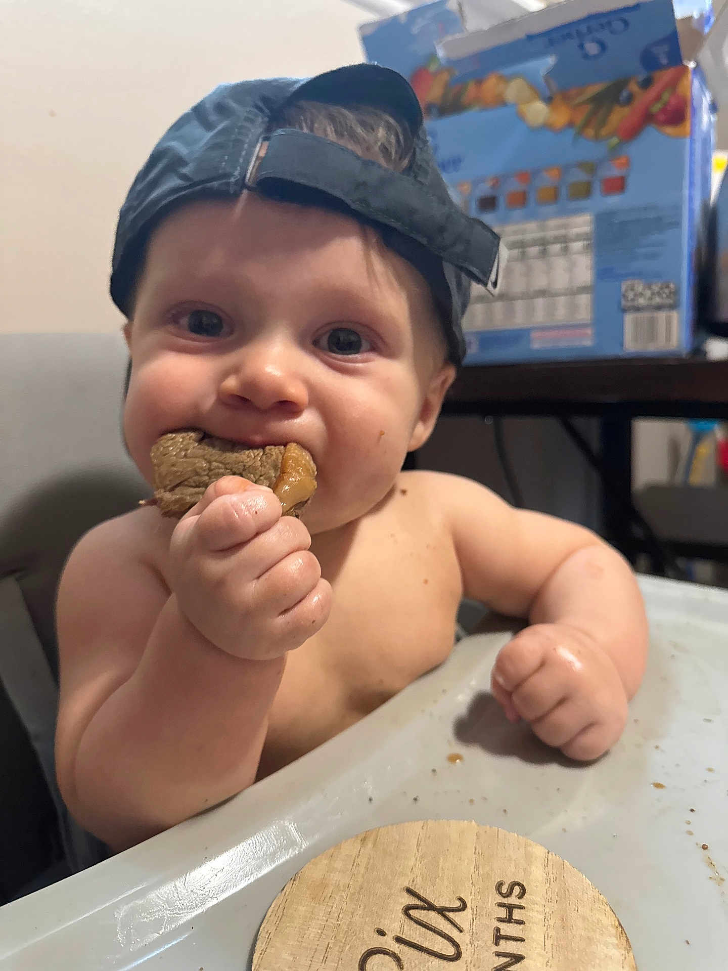 Cooper joined the competition — help win amazing prizes! baby, child, high_chair, food, meat, eating, cap, backwards_cap, hand, face, cheeks, milestone_marker, six_months, indoor, cute, chubby, table, person, skin, closeup