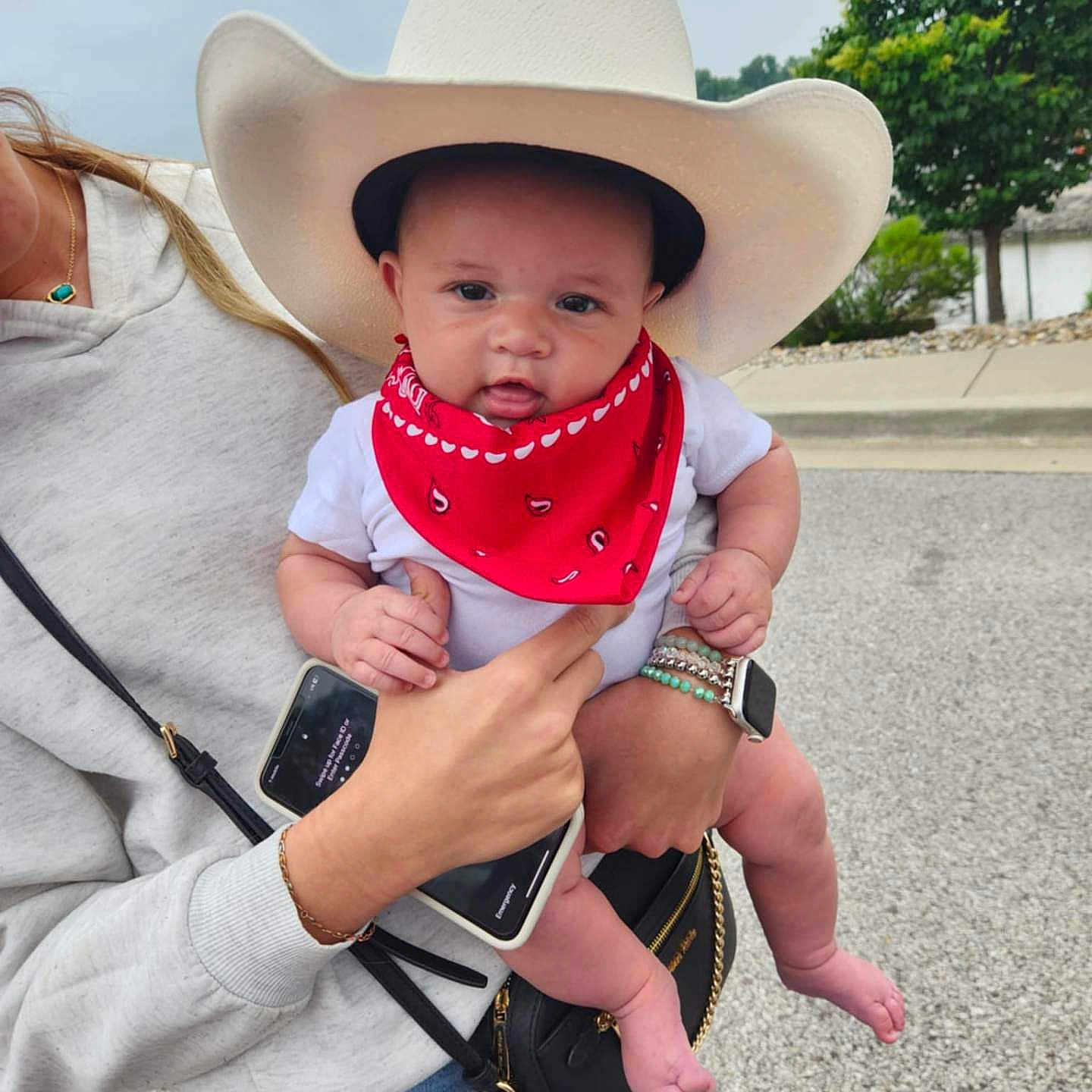Walker John is registered to the contest to win money with this photo: accessories, adult_hand, baby, bracelets, casual_clothing, child, cloudy_sky, cowboy_hat, cute, jeans, outdoor, pavement, person_holding_baby, portrait, red_bandana, small_feet, smartphone, smartwatch, tree, white_onesie