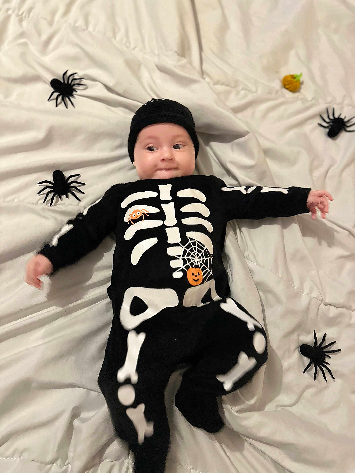 Sasha participe au concours pour gagner de l'argent avec cette photo : baby, child, costume, skeleton, black_clothing, white_blanket, toy_spiders, pumpkin, halloween, cute, lying_down, infant, smiling, headwear, indoors, playful, holiday, decorations, person, festive