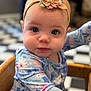 baby, child, headband, flower, blue_eyes, patterned_clothing, high_chair, wooden_furniture, close_up, indoor, curious_expression, face, infant, long_sleeves, colorful, soft_lighting, portrait, young_child, seated, cute