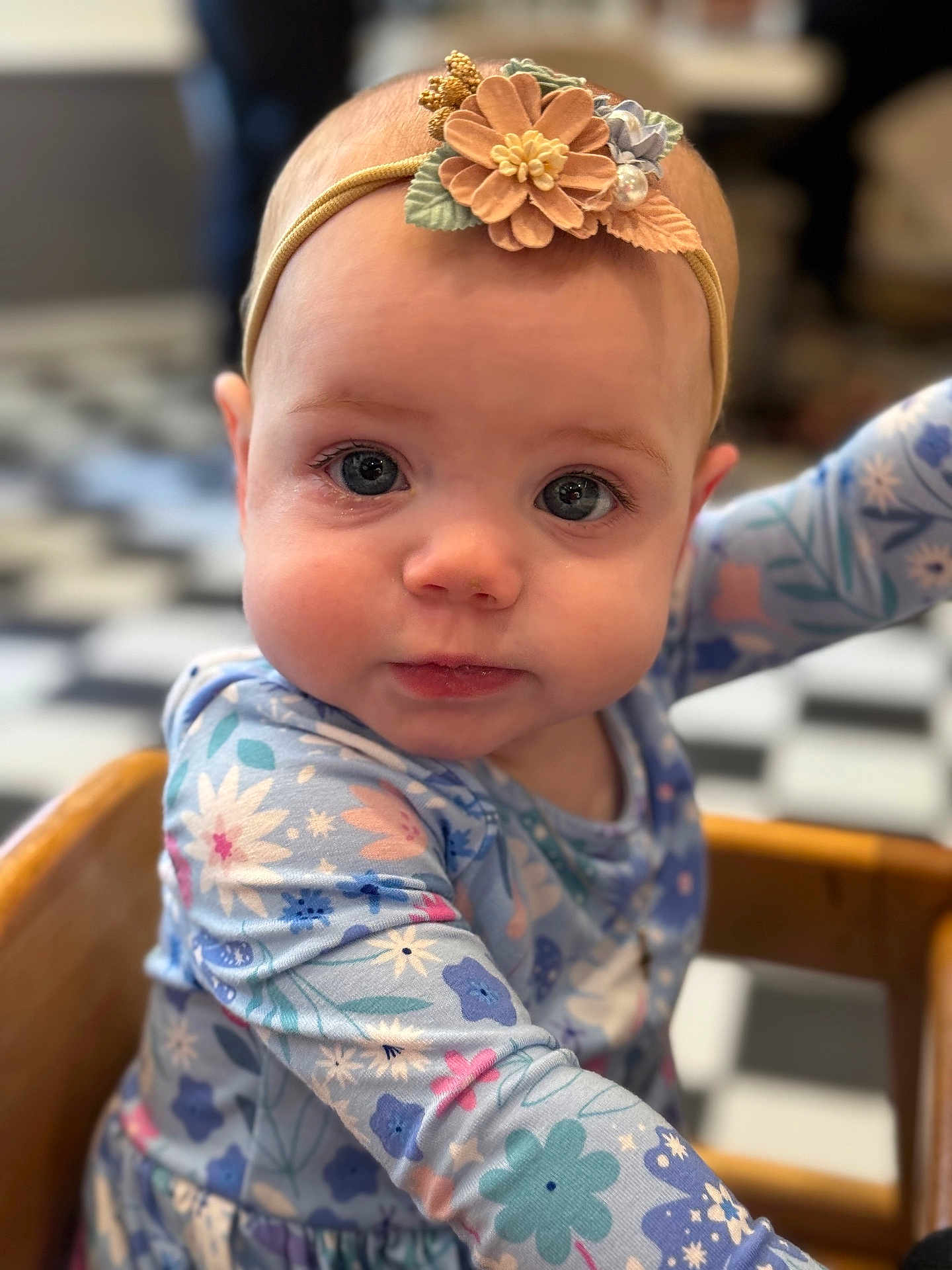 Amelia is registered to the contest to win money with this photo: baby, child, headband, flower, blue_eyes, patterned_clothing, high_chair, wooden_furniture, close_up, indoor, curious_expression, face, infant, long_sleeves, colorful, soft_lighting, portrait, young_child, seated, cute