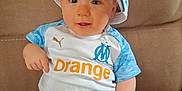 Tyrone participe au concours pour gagner de l'argent avec cette photo : baby, blue, cap, child, clothing, couch, cute, face, hat, indoor, jersey, orange_text, person, portrait, sitting, smiling, sofa, sportswear, white, young