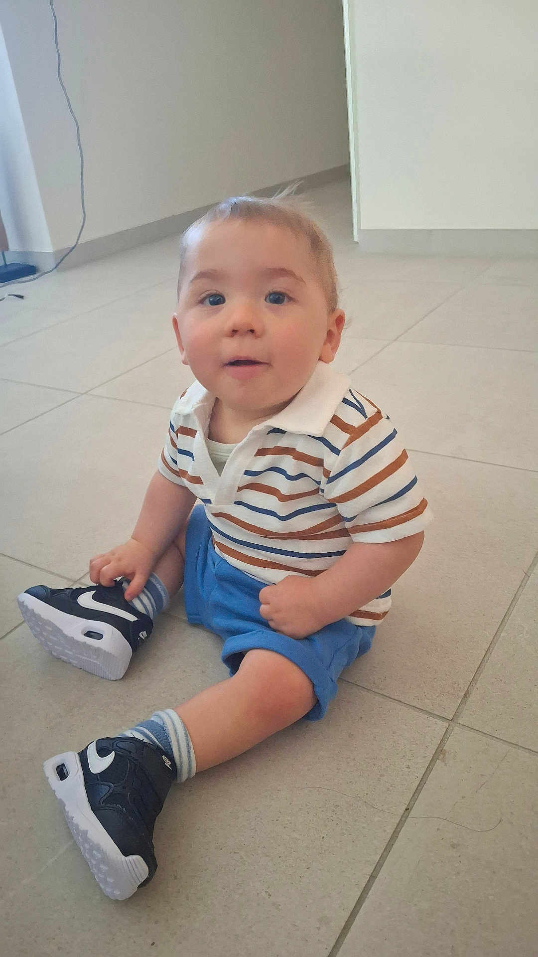 Tyrone participe au concours pour gagner de l'argent avec cette photo : baby, blue_shorts, casual_clothing, child, curious, cute, floor, home, indoor, light, neutral_background, person, portrait, sitting, sneakers, socks, striped_shirt, tiles, toddler, young_child