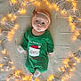 baby, infant, onesie, green_clothing, santa_claus, headband, red_flower, blanket, fairy_lights, warm_lighting, holiday_theme, lying_down, cute, face, indoors, cozy, christmas, child, festive, decorative_lights