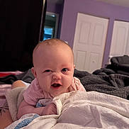 Monica is registered to the contest to win money with this photo: baby, child, infant, face, expression, blanket, bed, indoor, purple_wall, door, comfort, cute, playful, person, skin, clothing, toddler, room, smile, lying_down