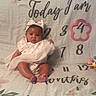 baby, infant, child, portrait, sitting, milestone_blanket, bow, dress, floral, indoors, bed, blanket, calendar, numbers, toy, feet, face, looking_at_camera, photography, family_milestone