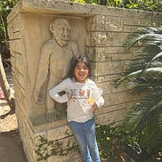 Divinity is registered to the contest to win money with this photo: child, girl, statue, stone, outdoor, sunlight, trees, leafy, wall, snacks, cup, smiling, casual_clothing, jeans, sandals, nature, daytime, plant, ivy, sidewalk