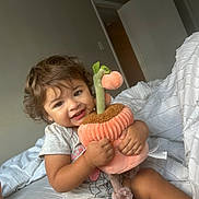 Evelyn is registered to the contest to win money with this photo: toddler, child, plush_toy, bed, blanket, smile, happy, curly_hair, indoors, person, clothing, cute, soft_toy, cuddling, young_child, toy, fabric, home, comfort, playful