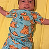 baby, infant, child, blue_clothing, cartoon_print, yellow_background, pacifier, lying_down, cute, face, arms_outstretched, sleepwear, patterned_clothes, indoors, soft_lighting, playful, expression, comfort, bed, rest