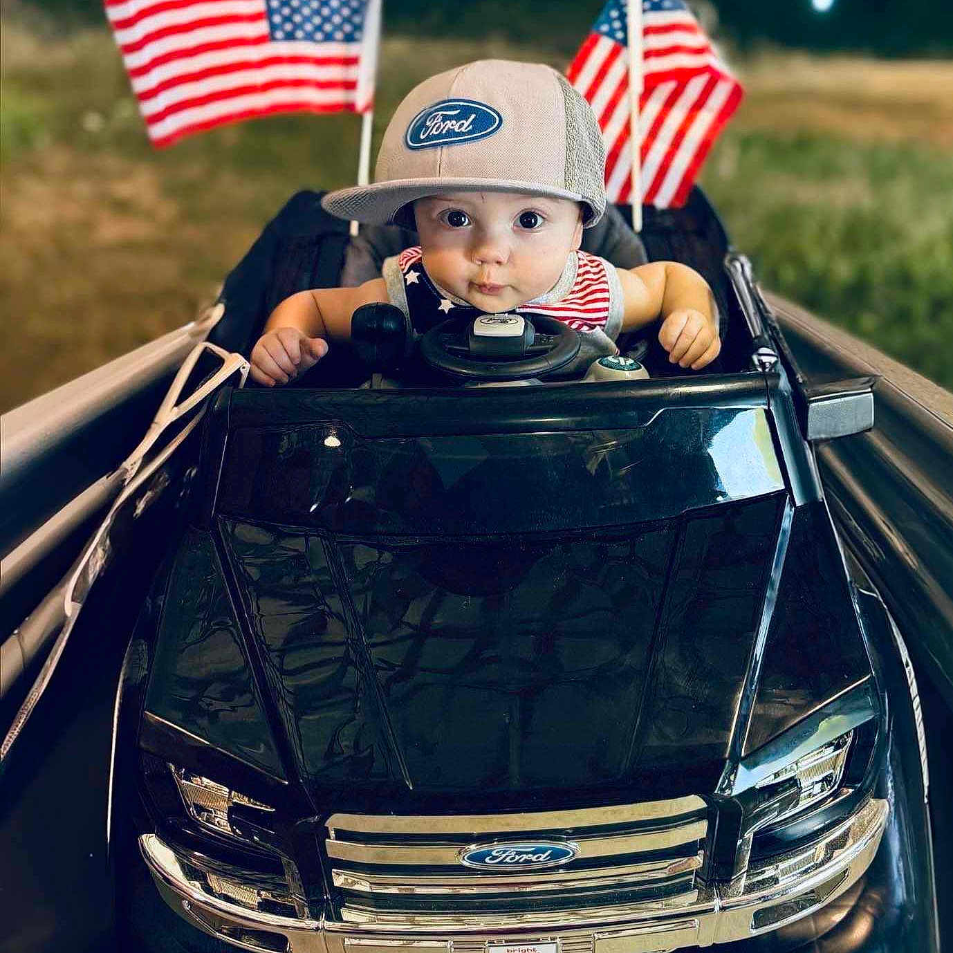 Ford is registered to the contest to win money with this photo: americanflag, baby, car, clothing, face, flag, fun, grass, hat, head, malaysiaflag, person, photography, plant, portrait, sitting, tire, transportation, vacation, vehicle