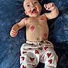 baby, infant, child, face, smile, kiss_marks, red_hearts, pants, white_clothing, blue_blanket, happy, joyful, open_mouth, raised_fists, lying_down, skin, cute, playful, portrait, indoors
