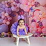child, baby, toddler, purple_bow, purple_tutu, balloons, pink_balloons, purple_balloons, butterflies, flowers, floral_arrangement, wooden_stool, studio_backdrop, birthday_theme, party_decor, fairy_lights, smiling, portrait, happy, decor