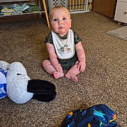 Wilder is registered to the contest to win money with this photo: baby, child, indoor, carpet, plush_toy, pelican, bib, camo_clothing, sitting, floor, blanket, dinosaur_pattern, curious, cute, young_child, furniture, basket, gate, door, room