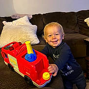 Cameron is registered to the contest to win money with this photo: toddler, child, toy, fire_truck, smiling, indoor, couch, pillow, blonde_hair, clothing, shoes, happy, furniture, person, play, living_room, fun, cute, young_child, colorful