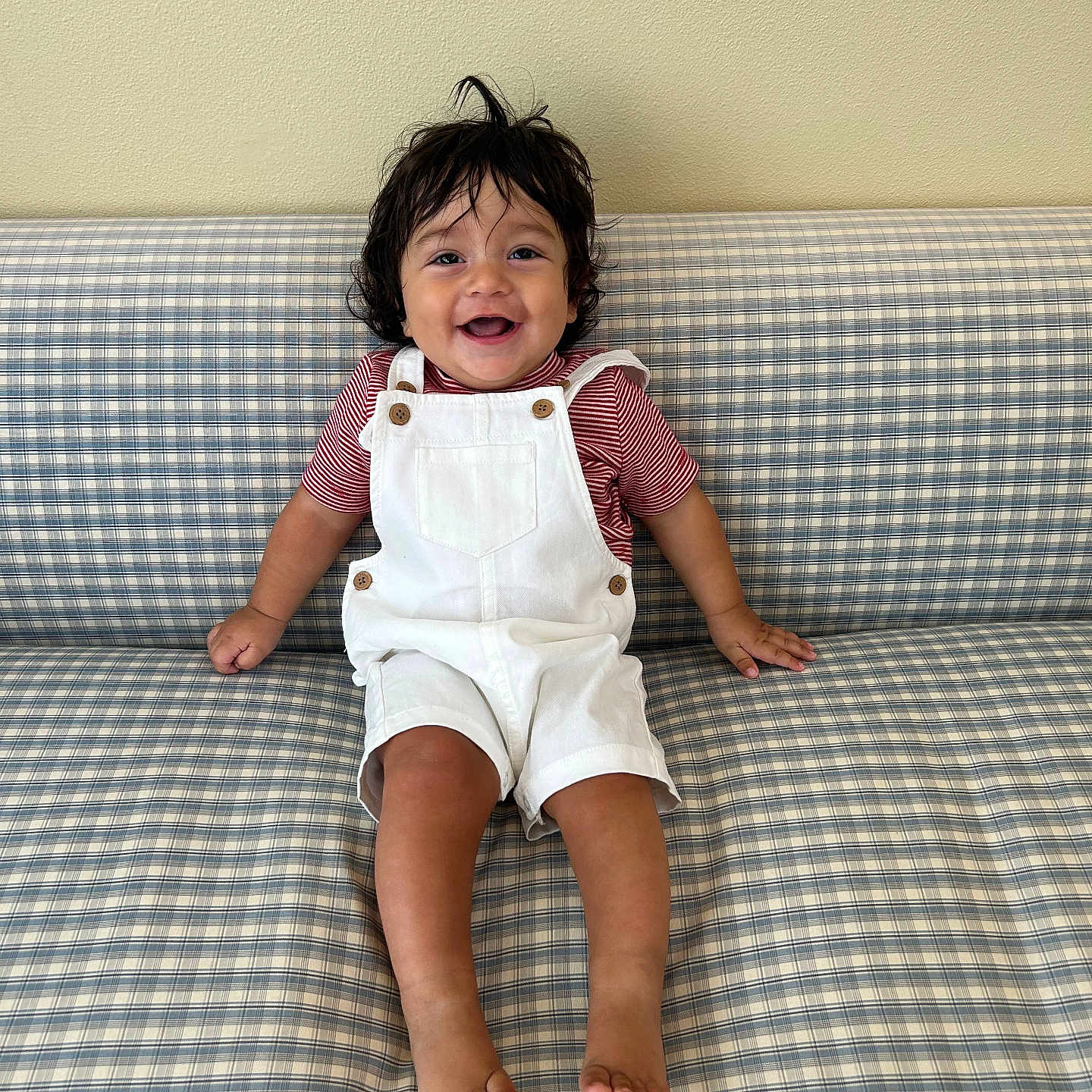 Sebastian is registered to the contest to win money with this photo: toddler, child, smiling, happy, sitting, couch, checkered_pattern, overalls, striped_shirt, barefoot, indoor, person, hair, face, joy, cute, young_child, plaid, furniture, casual_clothing