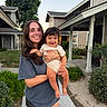 woman, baby, child, smile, outdoor, suburban, house, greenery, happy, holding, person, casual_clothing, shorts, tshirt, sidewalk, daylight, portrait, family, cute, nature