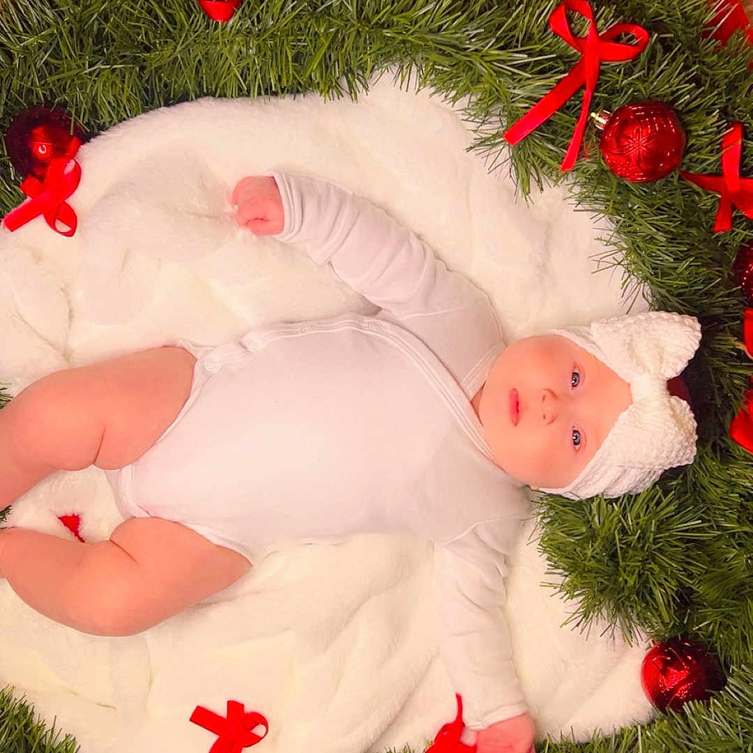 Emely is registered to the contest to win money with this photo: baby, blanket, celebration, christmas_ornaments, chubby_cheeks, cute, decorations, festive, greenery, holiday, indoors, infant, knitted_hat, lying_down, portrait, red_background, red_bows, soft_texture, white_onesie, wreath