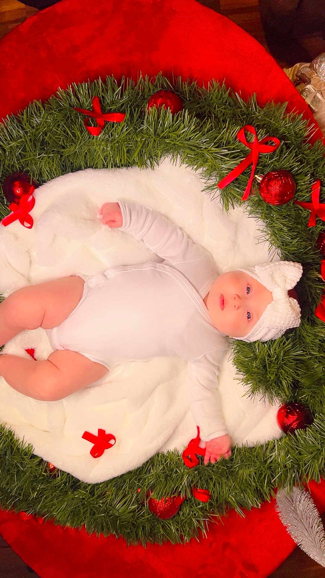 Emely is registered to the contest to win money with this photo: baby, infant, white_onesie, knitted_hat, blanket, wreath, greenery, red_bows, christmas_ornaments, holiday, festive, red_background, cute, chubby_cheeks, lying_down, indoors, soft_texture, decorations, celebration, portrait