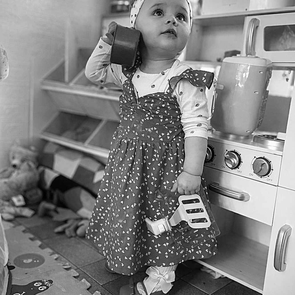 Gloria a rejoint le concours — aidez-le/la à gagner de superbes lots ! toddler, child, toy, dress, headband, kitchen, playroom, floor_mat, pretend_play, monochrome, cooking_toys, footwear, innocence, curiosity, indoor, young_child, play_kitchen, holding, looking_up, cute