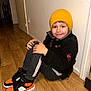 child, yellow_hat, black_jacket, smiling, indoor, wooden_floor, sneakers, orange, black, gray_pants, wall, floor, person, casual_clothing, happy, young, seated, cozy, fashion, portrait
