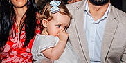 Alma a rejoint le concours — aidez-le/la à gagner de superbes lots ! toddler, child, thumb_sucking, white_dress, blue_bow, adult, woman, man, beige_suit, red_patterned_clothing, portrait, indoor, family, people, close_up, cute, shy, hair_bow, casual, event
