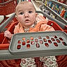 baby, shopping_cart, child, store, aisle, books, clothing, hoodie, pants, floral_pattern, red_cart, person, indoor, retail, curious, wide_eyes, floor, socks, hand, face