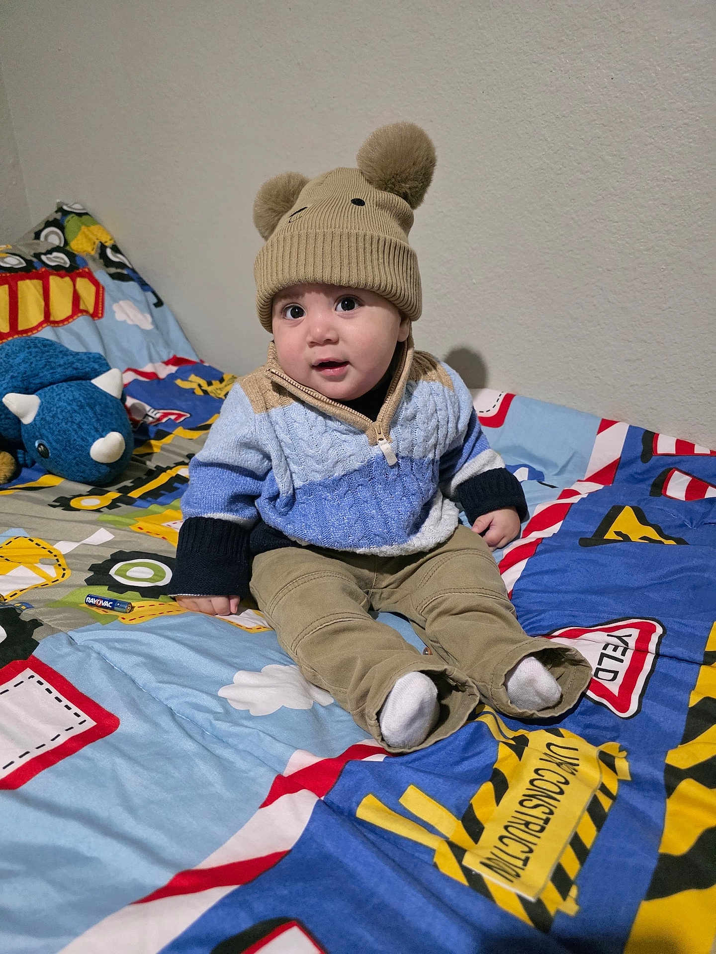 Jared is registered to the contest to win money with this photo: baby, child, hat, knitwear, sweater, pants, socks, bedspread, toy, plush, dinosaur, indoor, child_bedroom, cute, smiling, seated, colorful, cozy, playful, portrait