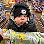 baby, child, shopping_cart, blanket, black_jacket, beanie, smiley_face, face, eyes, hand, store, aisle, shelf, toys, floor, person, winter_clothing, curious, indoor, shopping