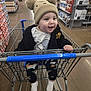 Jared is registered to the contest to win money with this photo: baby, child, shopping_cart, grocery_store, hat, beanie, clothing, black_jacket, smiling, happy, indoor, aisle, products, socks, pants, person, retail, floor, hand, face