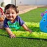 Joseph is registered to the contest to win money with this photo: toddler, child, grass, outdoor, smile, green_mat, blue_bunny_bag, gift_bag, red_bow, curly_hair, short_sleeves, playful, daylight, sunlight, happy, cute, toy, crawling, casual_clothing, park