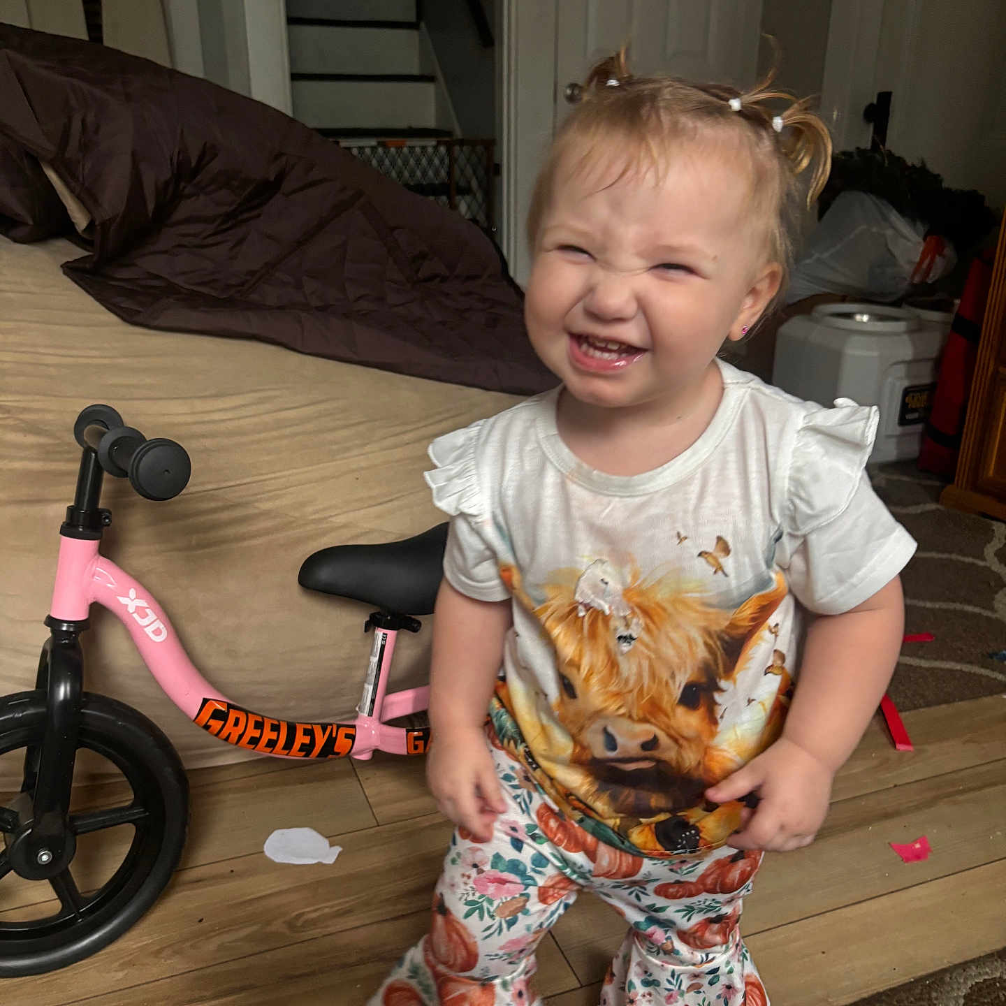 Braelyn is registered to the contest to win money with this photo: balance_bike, bell_bottoms, child, clothing, cow_print, door, floral_pants, happy, home, indoor, person, pigtails, pink_bike, playful, rug, smiling, stairs, toddler, white_shirt, wooden_floor