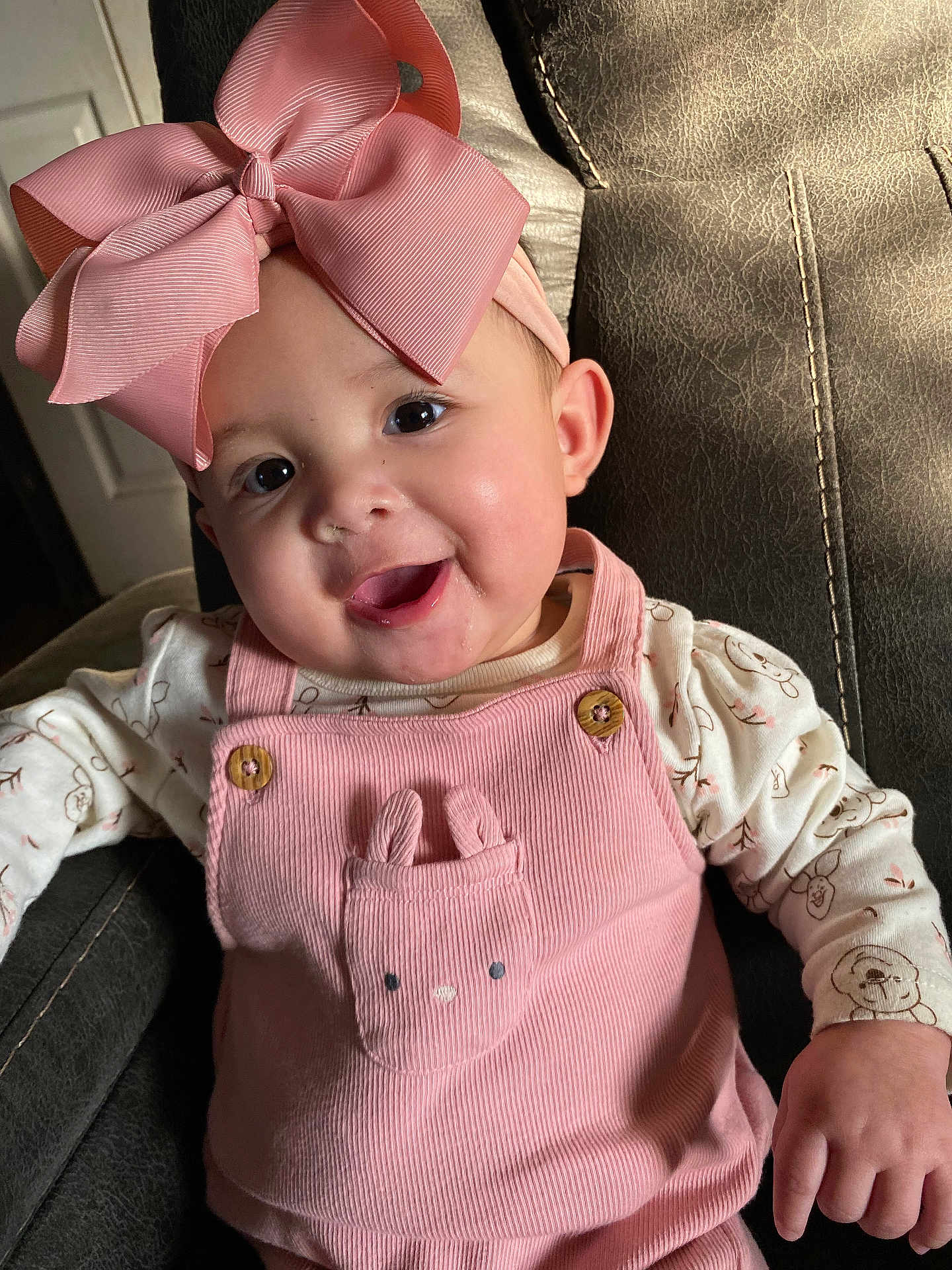 Naomi is registered to the contest to win money with this photo: baby, child, pink_bow, overalls, bunny_pocket, smiling, happy, cute, indoor, leather_chair, clothing, portrait, infant, headband, face, person, toddler, apparel, seated, adorable