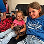 child, smiling, puppy, dog, couch, jeans, hoodie, indoor, happy, black_leather, pink_shirt, blue_hoodie, hair_accessory, living_room, lamp, pillow, family, casual, cute, friendship