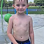 Zayden is registered to the contest to win money with this photo: child, boy, wet_hair, swim_trunks, outdoor, splash_pad, water_play, summer, sunny, smiling, young, recreation, fun, blue, skin, standing, background, fence, trees, park