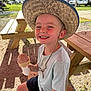 child, boy, smiling, hat, straw_hat, ice_cream, cone, picnic_bench, outdoor, sunlight, summer, happy, casual_clothing, shorts, tshirt, grass, trees, park, daytime, recreation