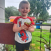 Zayden is registered to the contest to win money with this photo: child, boy, plant, potted_plant, smiling, outdoor, porch, grass, tree, house, shirt, spiderman, clothing, fence, railing, greenery, daylight, casual, happy, person