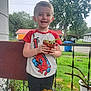 child, boy, plant, potted_plant, smiling, outdoor, porch, grass, tree, house, shirt, spiderman, clothing, fence, railing, greenery, daylight, casual, happy, person