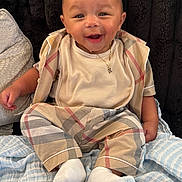 Khari is registered to the contest to win money with this photo: baby, smiling, infant, pajamas, white_socks, blanket, plaid, child, cute, portrait, happy, sitting, face, clothing, indoor, cozy, necklace, soft_toys, comfort, background